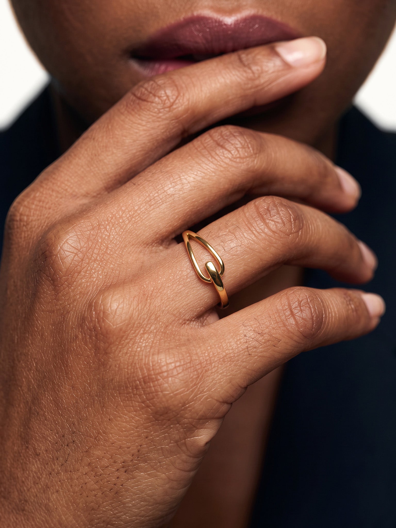Held Ring – worn with the fingers resting at the chin, close-up, gold, dark blazer, ivory studio background