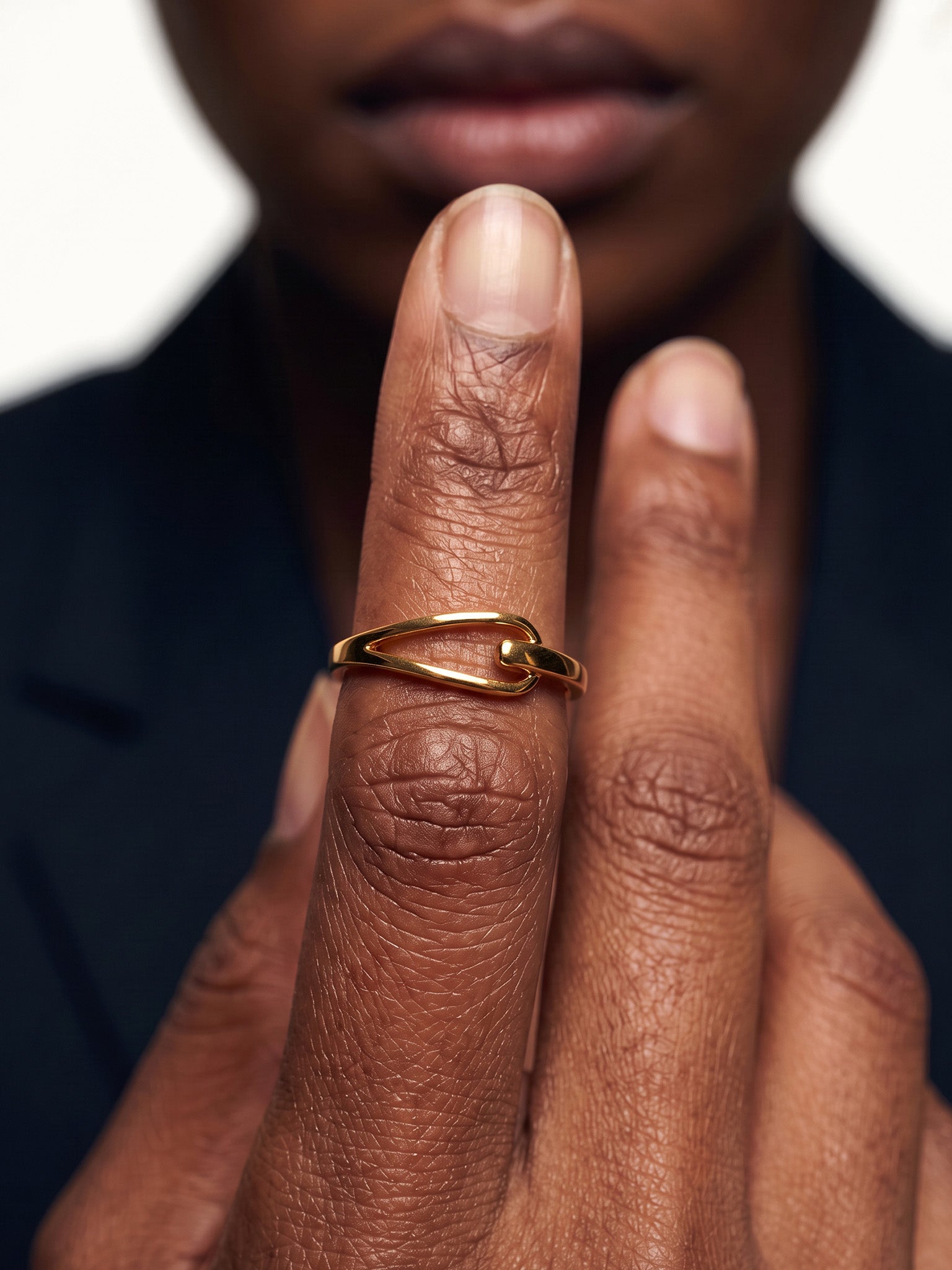 Held Ring – worn on fingers raised in front of the lips, close-up, gold, dark blazer, ivory studio background