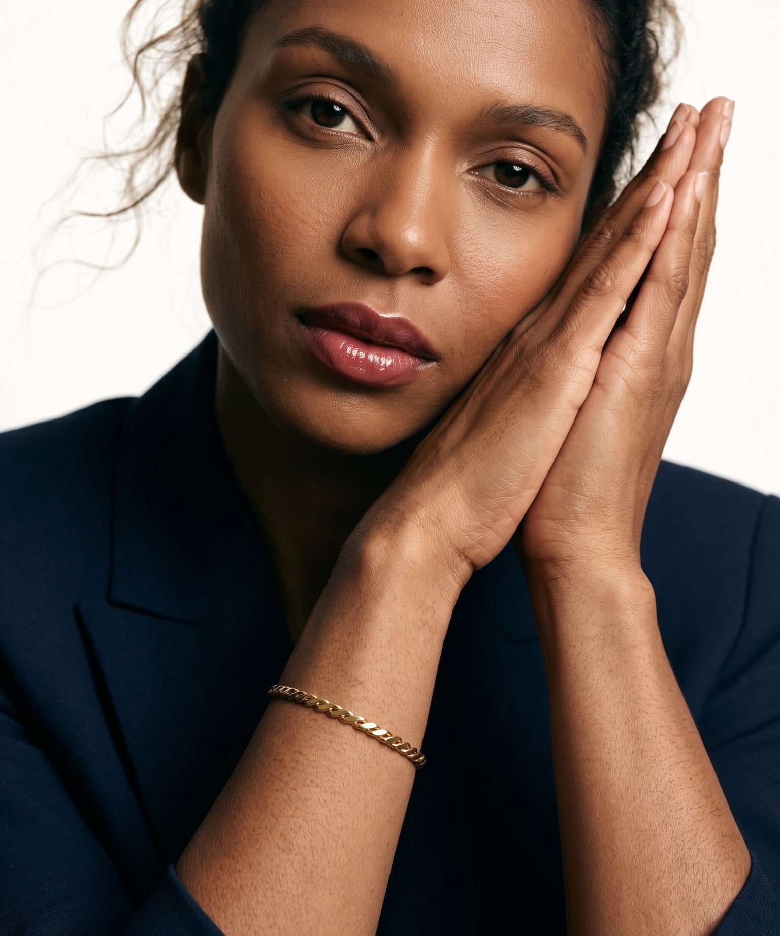 Kept Season Bangle – worn on the wrist, hands pressed together beside the face, gold, dark blazer, ivory studio background