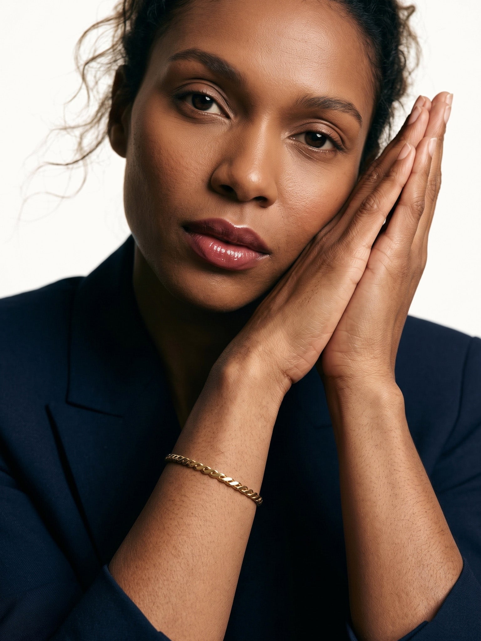 Kept Season Bangle – worn on the wrist, hands pressed together beside the face, gold, dark blazer, ivory studio background