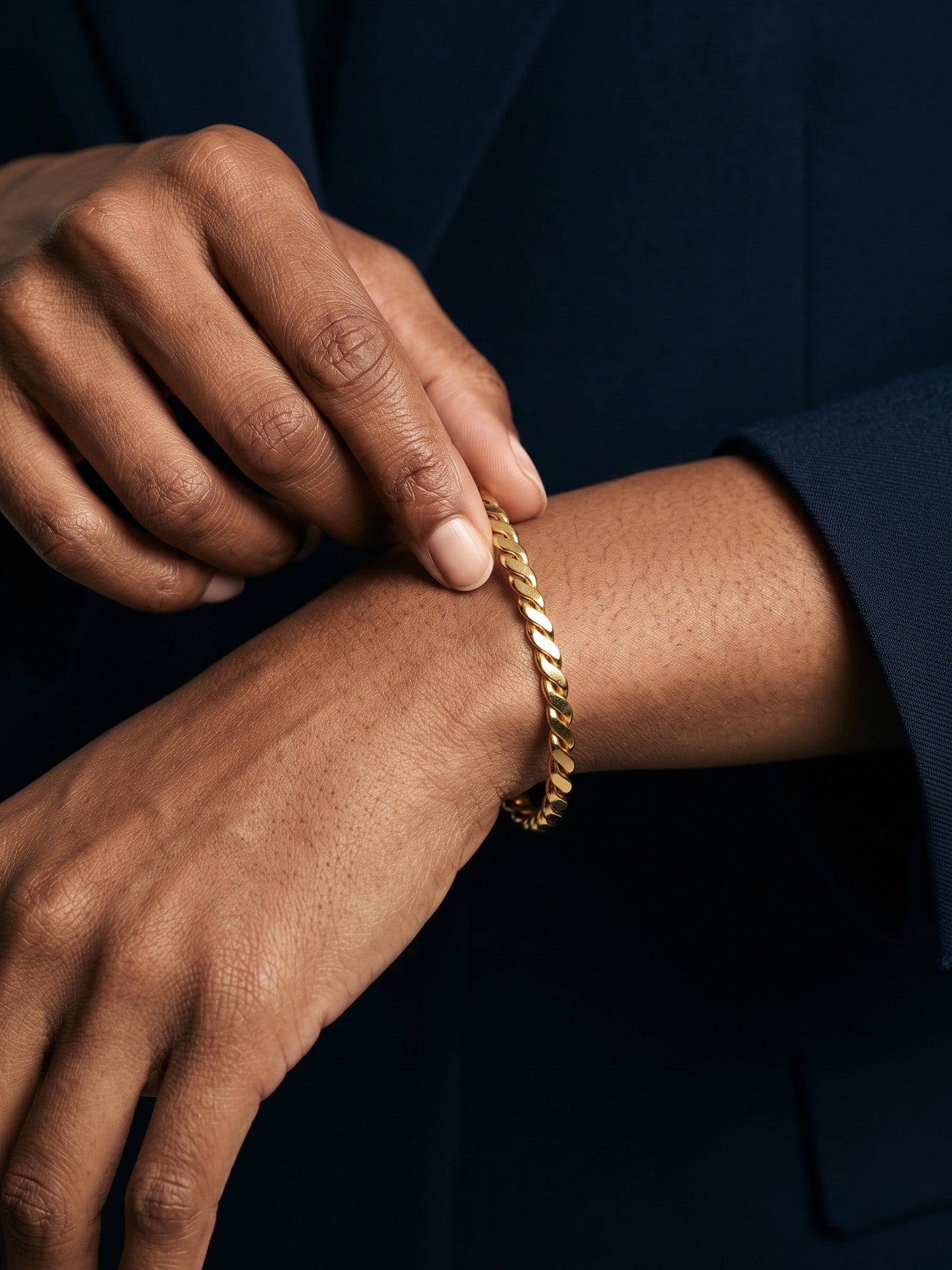 Kept Season Bangle – worn on the wrist, close-up, one hand adjusting the bangle, gold, dark blazer, ivory studio background