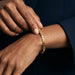 Kept Season Bangle – worn on the wrist, close-up, one hand adjusting the bangle, gold, dark blazer, ivory studio background