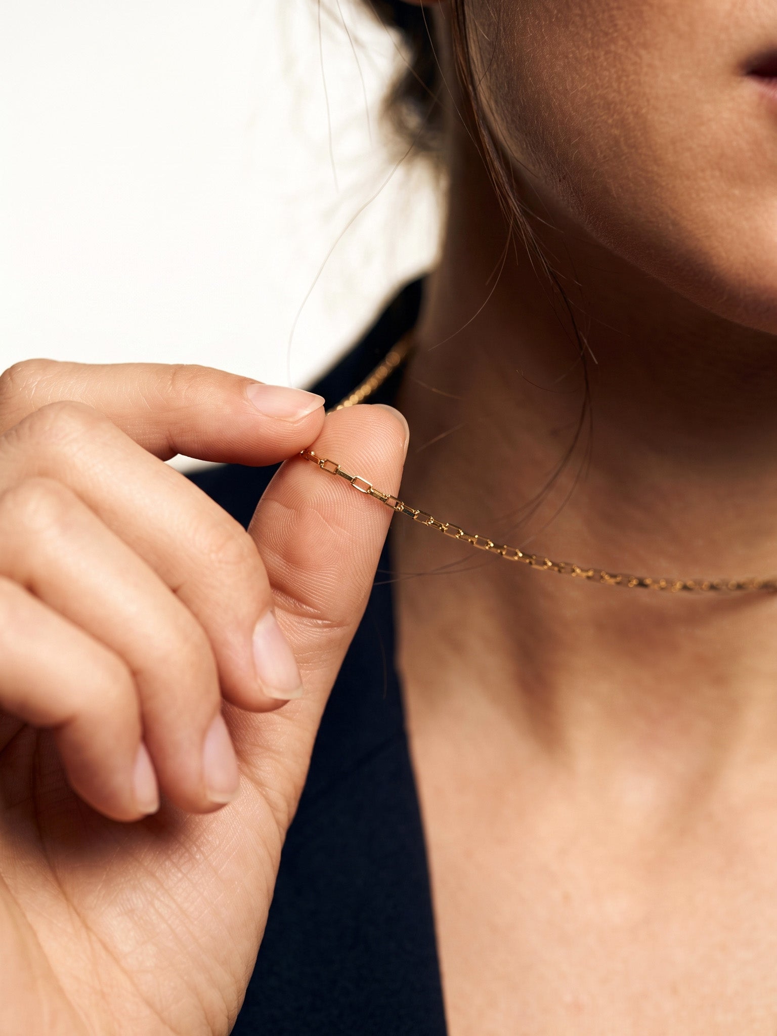 Kindred Link Necklace – fingers pinching the chain near the neck, close crop, gold, navy blazer, ivory studio background