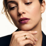 Mercies Ring – worn on a hand resting beneath the chin, gold, dark blazer, ivory studio background