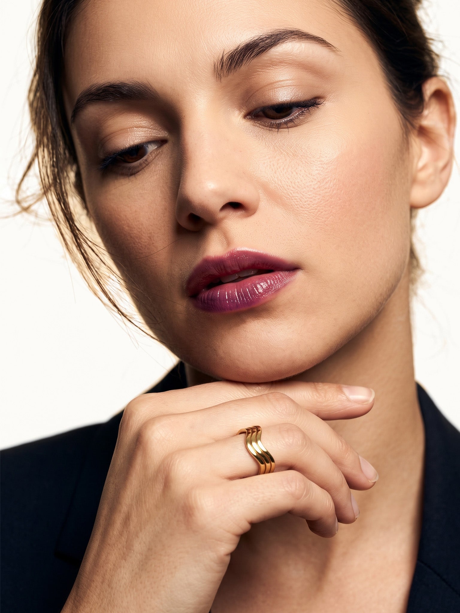 Mercies Ring – worn on a hand resting beneath the chin, gold, dark blazer, ivory studio background