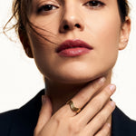 Mercies Ring – worn with the hand resting at the neck, gold, dark blazer, ivory studio background
