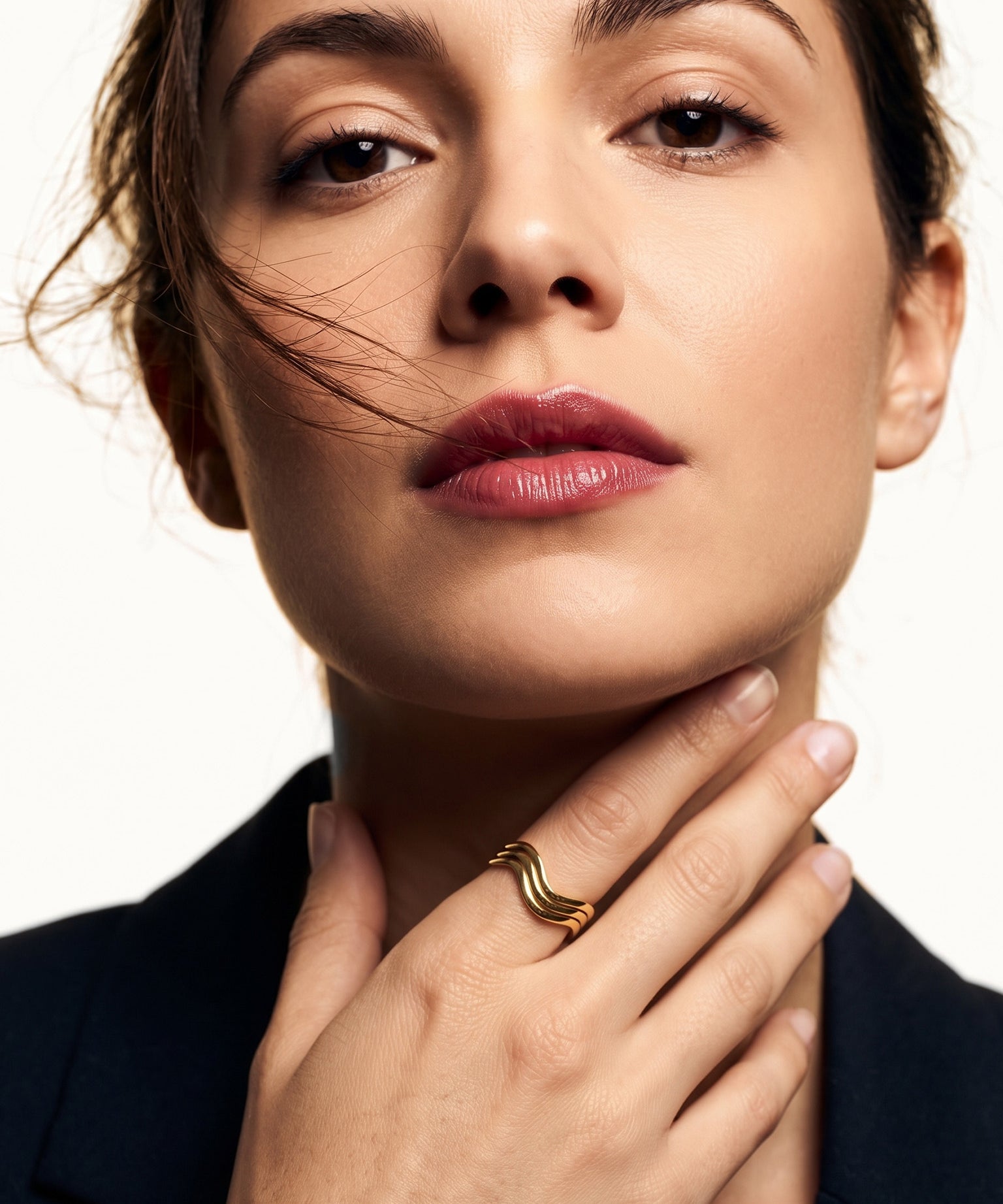 Mercies Ring – worn with the hand resting at the neck, gold, dark blazer, ivory studio background