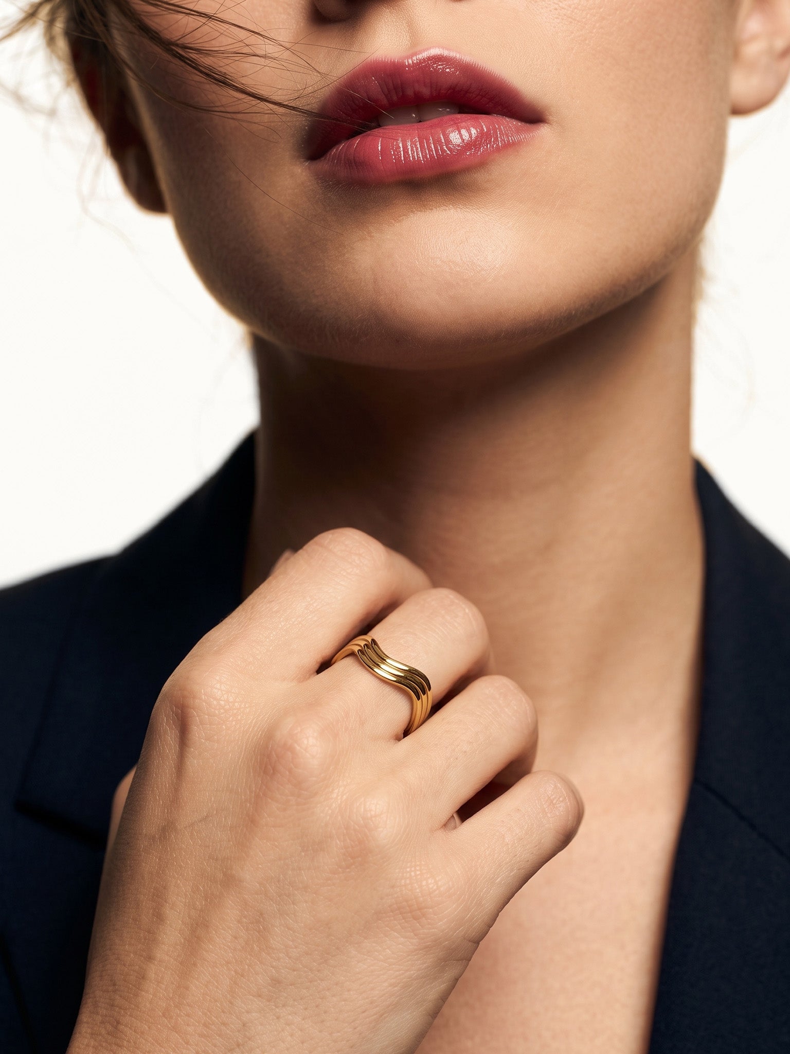 Mercies Ring – worn on a closed hand at the collarbone, gold, dark blazer, ivory studio background