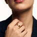 Mercies Ring – worn on a closed hand at the collarbone, gold, dark blazer, ivory studio background