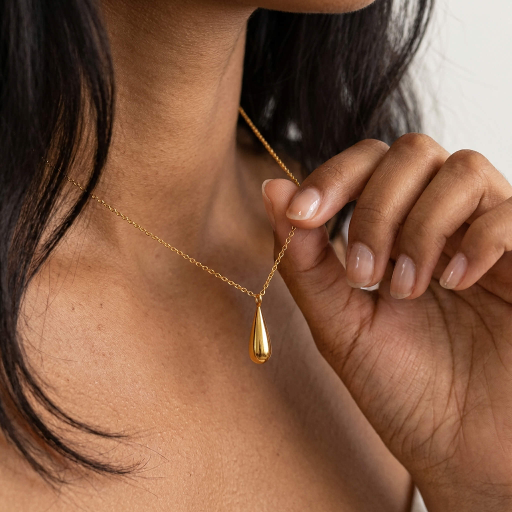 Mercy Teardrop Necklace – worn at the neck, fingers holding the teardrop, gold, bare shoulders