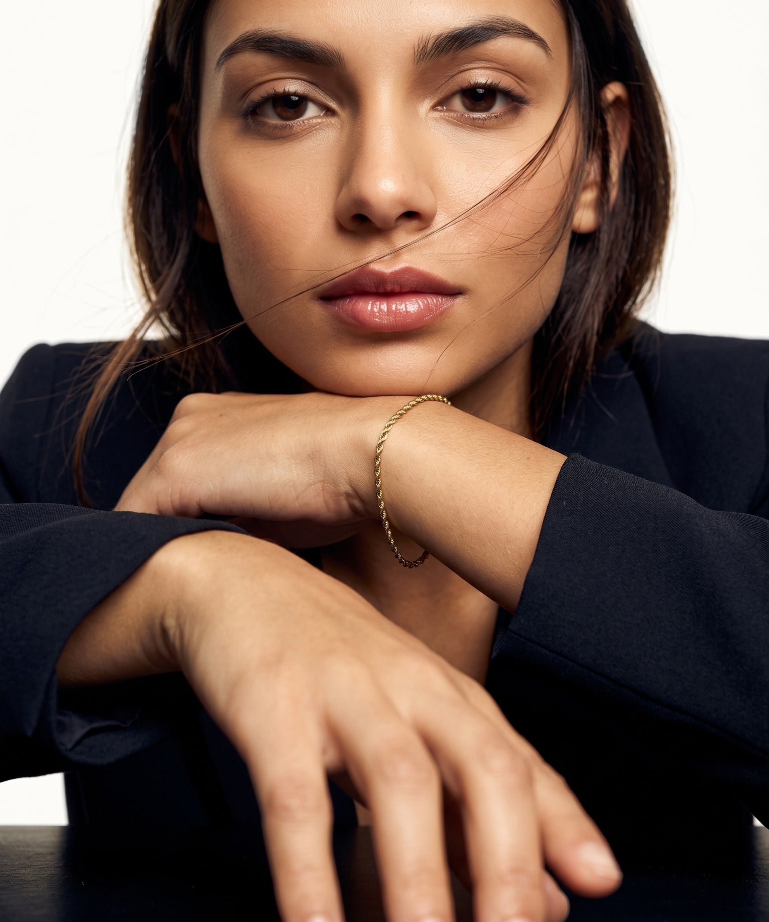Morning Light Twist Bracelet – worn on the wrist, chin resting on folded arms, gold, dark blazer, ivory studio background