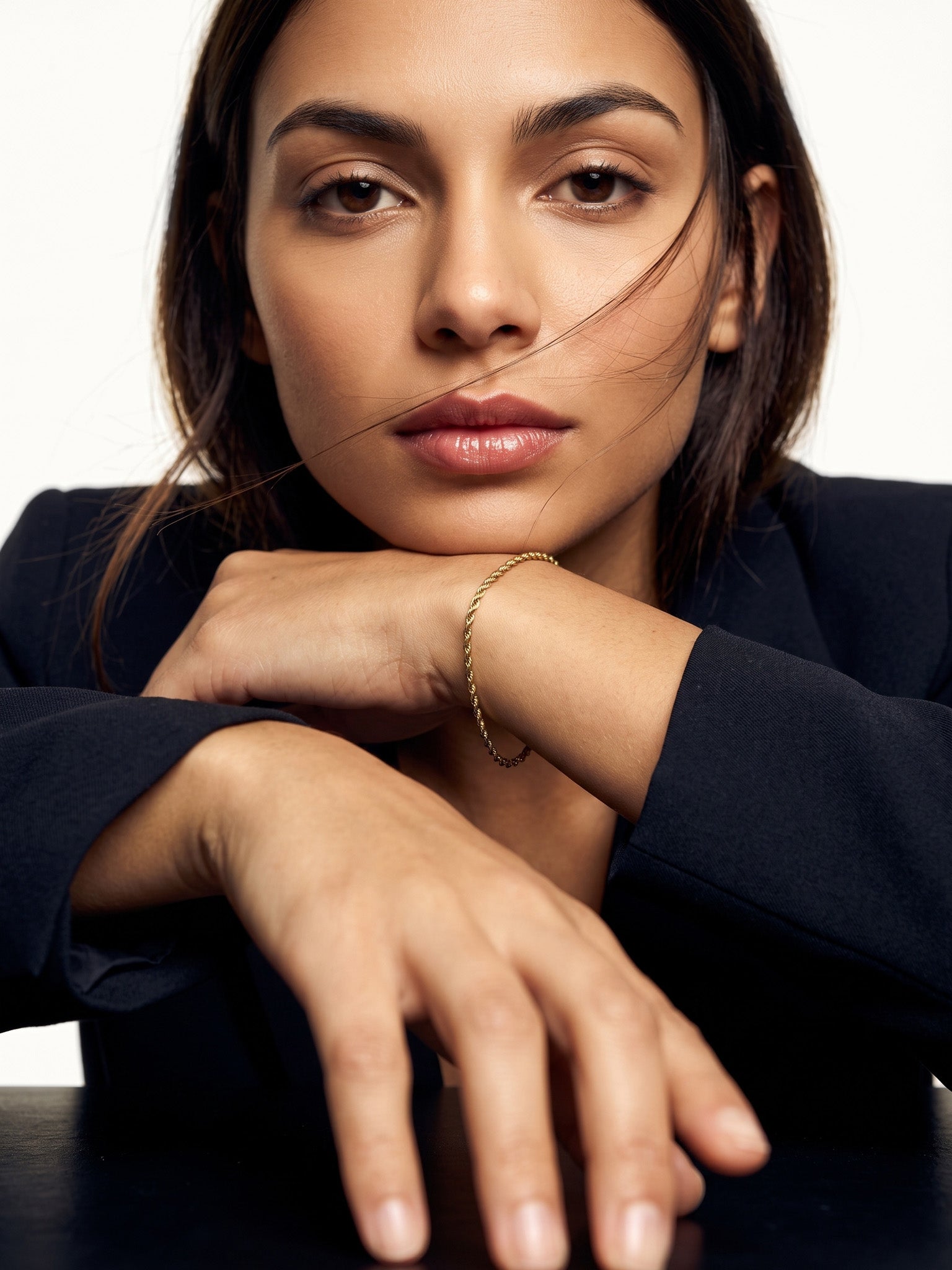 Morning Light Twist Bracelet – worn on the wrist, chin resting on folded arms, gold, dark blazer, ivory studio background