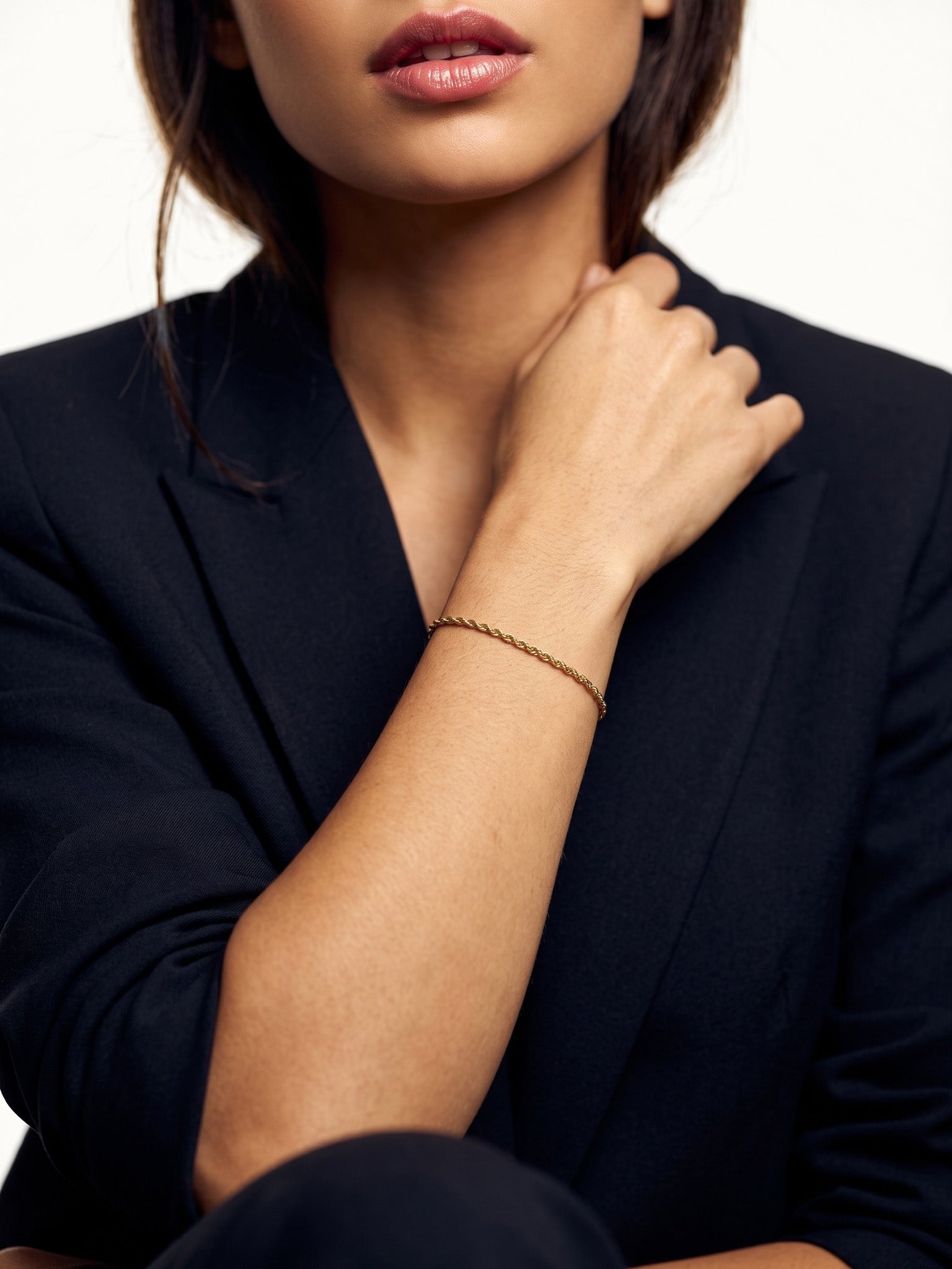 Morning Light Twist Bracelet – worn on the wrist, hand raised to the neck, gold, dark blazer, ivory studio background