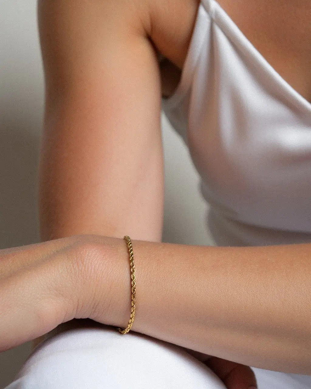 Morning Light Twist Bracelet – worn on the wrist, arm resting on the knee, gold, white satin top