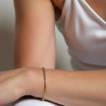 Morning Light Twist Bracelet – worn on the wrist, arm resting on the knee, gold, white satin top