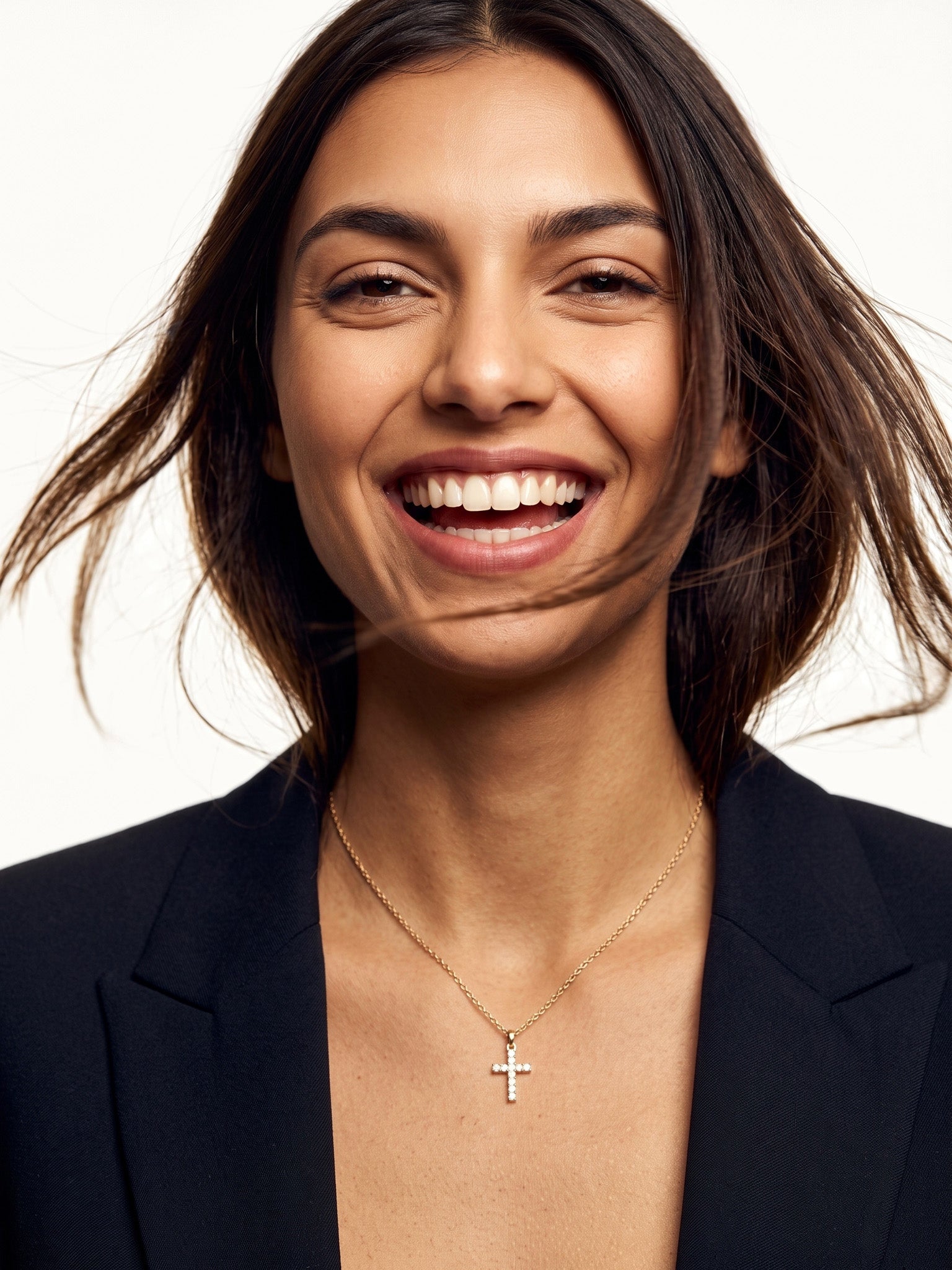 Radiant Cross Necklace – worn on the neck, laughing at the camera, gold cross with clear stones on a fine chain, dark blazer