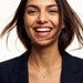 Radiant Cross Necklace – worn on the neck, laughing at the camera, gold cross with clear stones on a fine chain, dark blazer