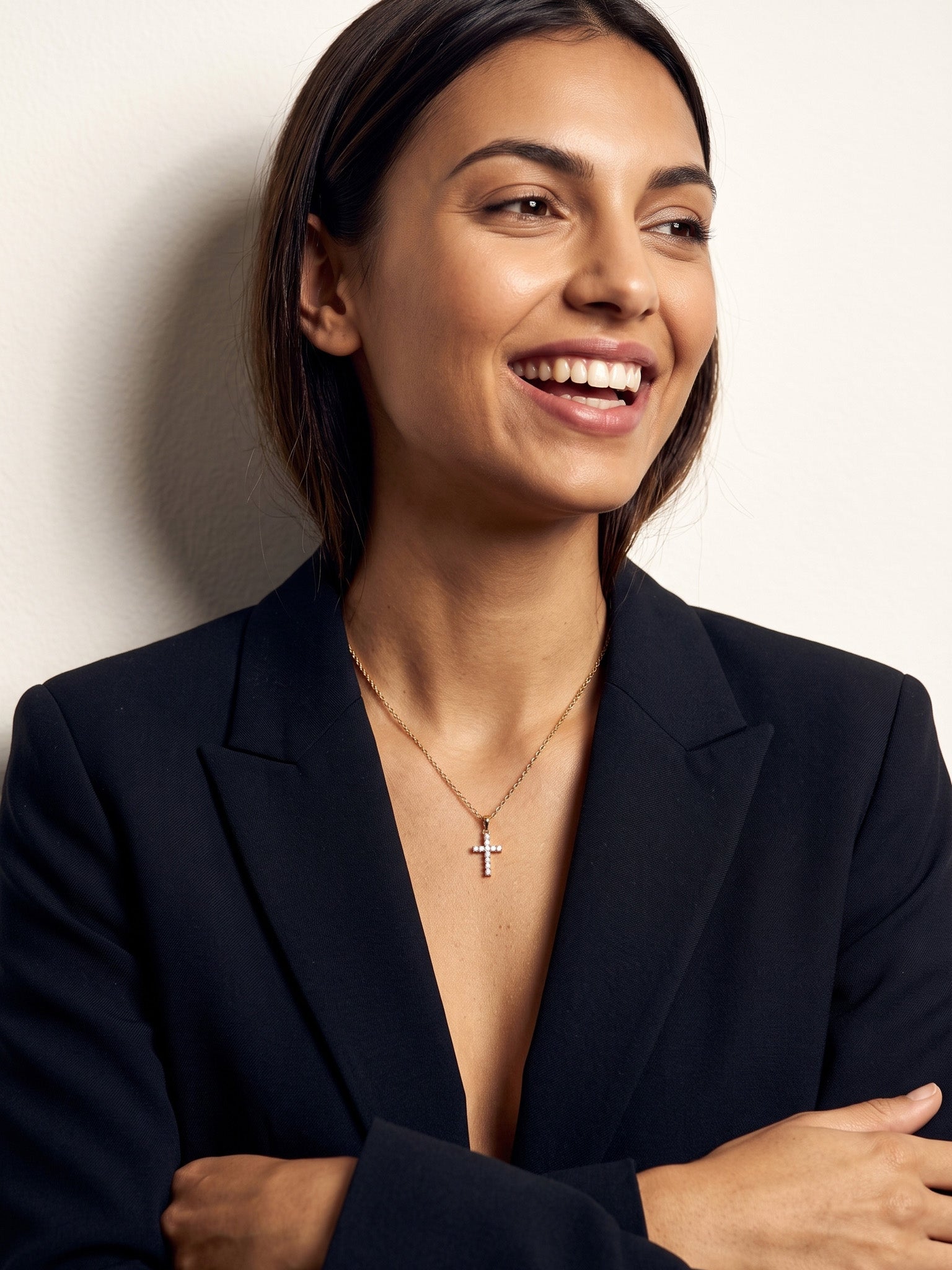 Radiant Cross Necklace – worn on the neck, smiling and looking aside with arms crossed, gold cross with clear stones on a fine chain