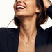 Radiant Cross Necklace – worn on the neck, laughing with a hand in the hair, gold cross with clear stones on a fine chain, dark blazer