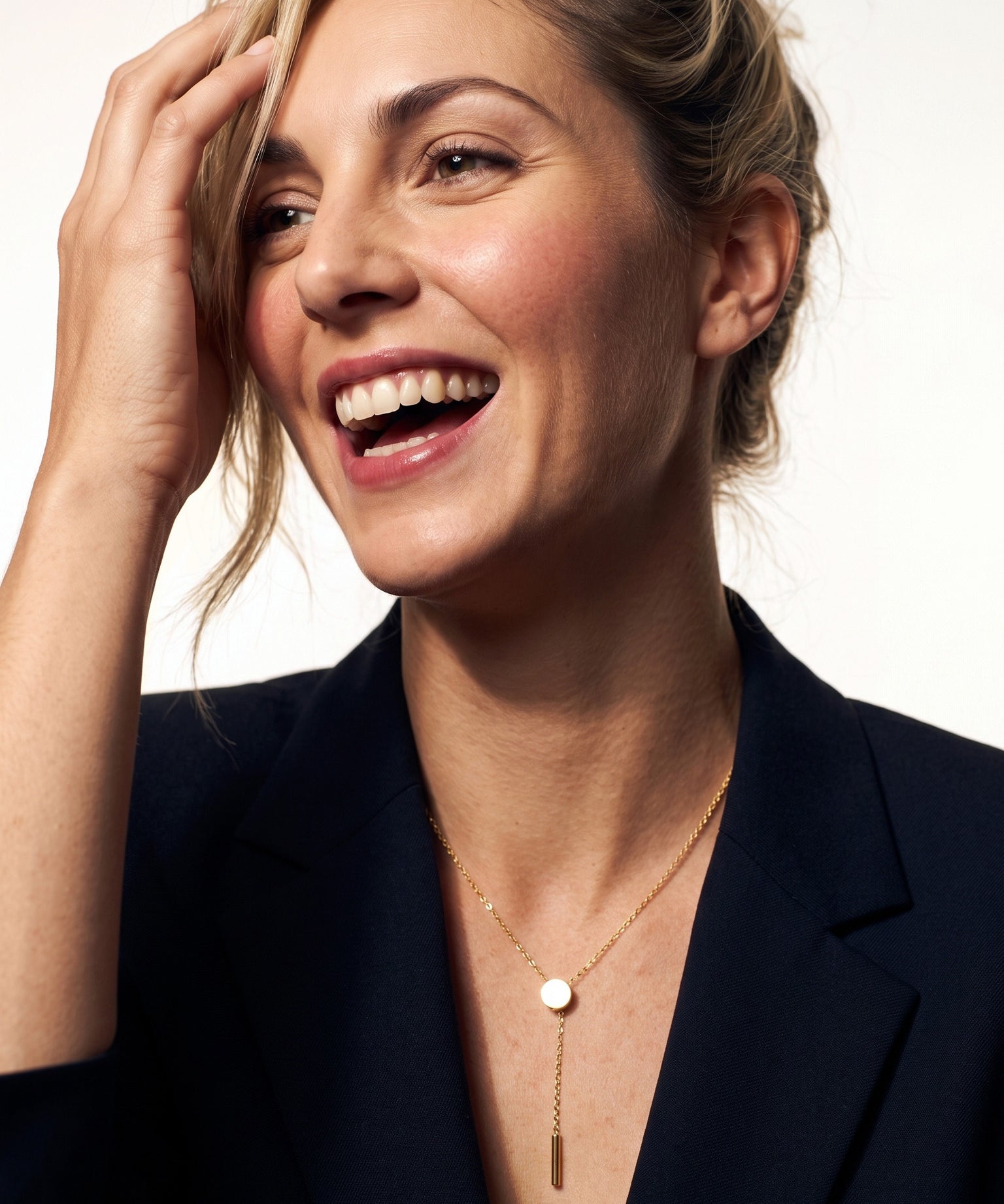 Rising Lariat Necklace – gold lariat with disc slider and bar drop worn by a laughing model with one hand in her hair, navy blazer
