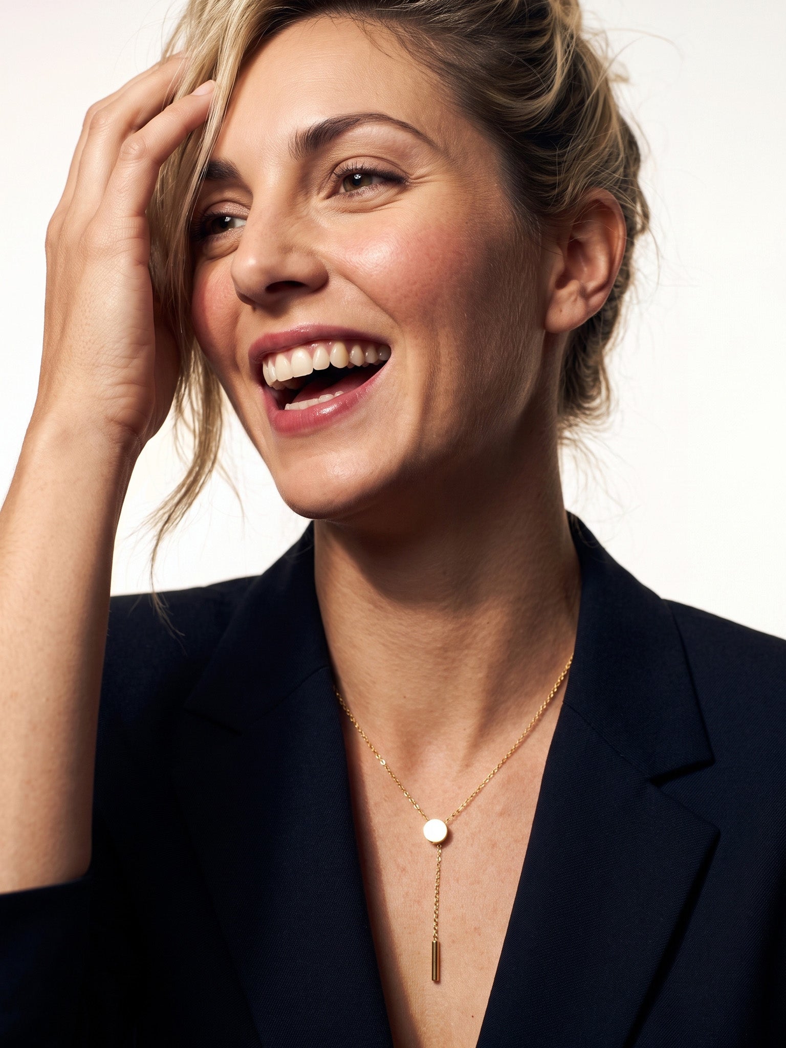 Rising Lariat Necklace – gold lariat with disc slider and bar drop worn by a laughing model with one hand in her hair, navy blazer