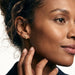 Rooted Cross Studs – worn in the ear, side profile with a hand at the jaw, gold cross stud, curly hair up