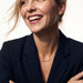 Signature Name Necklace – name "Jessica" in script at the collarbone, woman laughing and looking to one side, arms folded, gold, dark blazer, ivory studio background