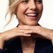 Signature Name Necklace – name "Jessica" in script at the collarbone, woman laughing with both hands under her chin, gold, dark blazer, ivory studio background