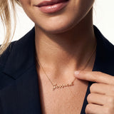 Signature Name Necklace – close view of the name "Jessica" in script on a fine chain, fingertips resting beside it, gold, dark blazer, ivory studio background