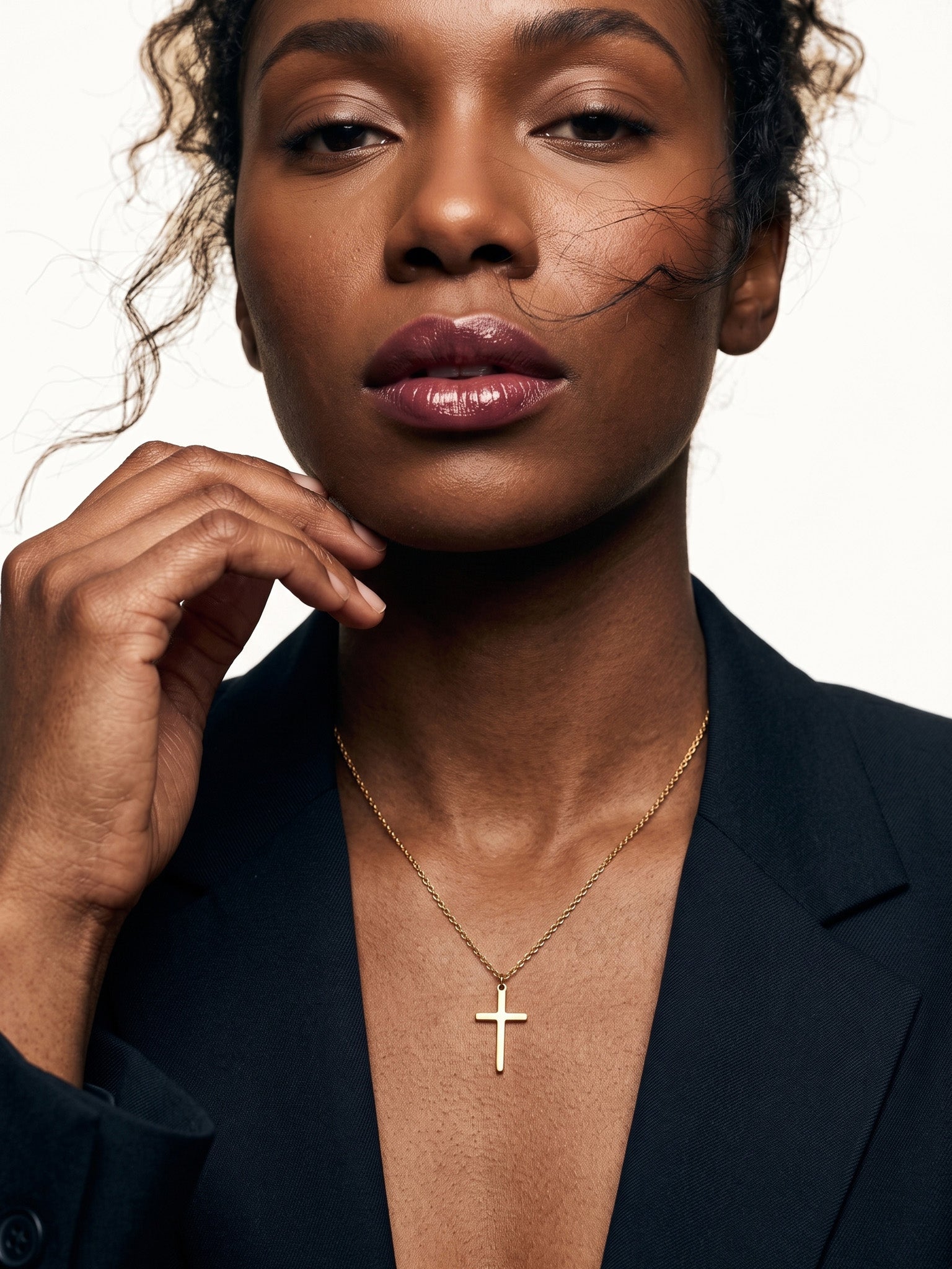 Steadfast Cross Necklace – worn on the neck, one hand at the jaw, gold, dark blazer, ivory studio background