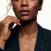 Steadfast Cross Necklace – worn on the neck, one hand at the jaw, gold, dark blazer, ivory studio background