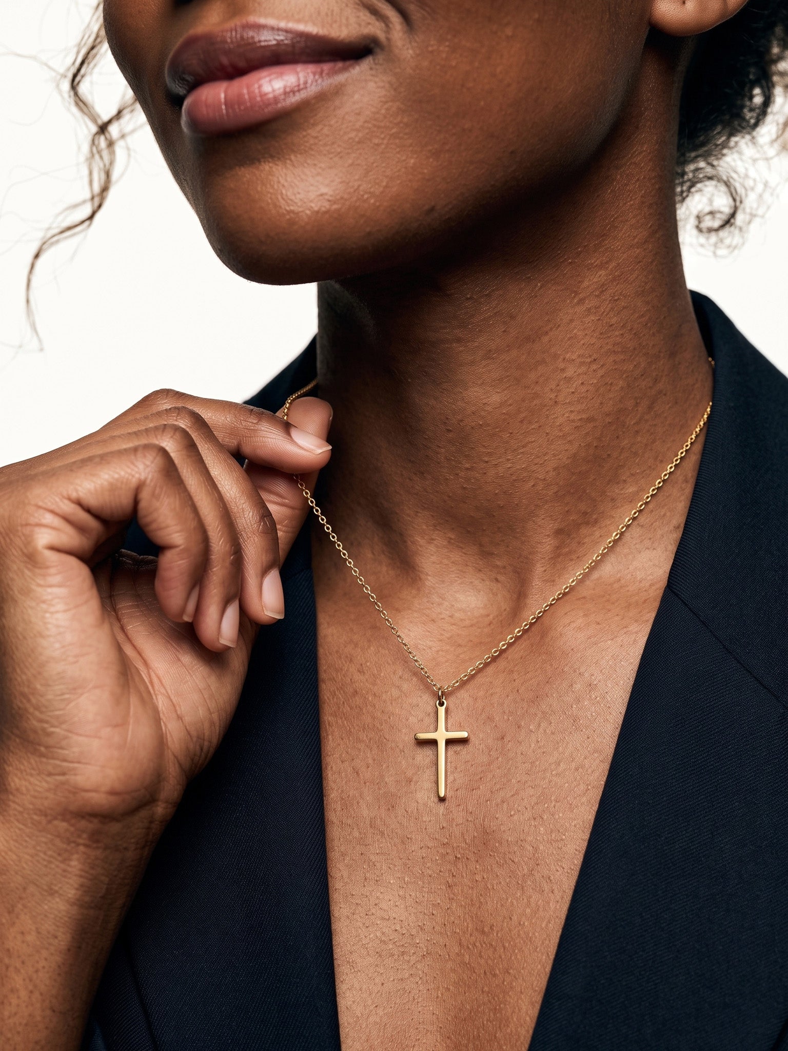 Steadfast Cross Necklace – worn on the neck, hand lifting the chain, close crop, gold, dark blazer, ivory studio background