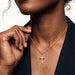 Steadfast Cross Necklace – worn on the neck, hand lifting the chain, close crop, gold, dark blazer, ivory studio background