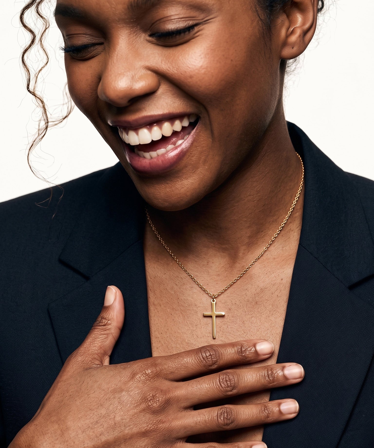 Steadfast Cross Necklace – worn on the neck, laughing, hand resting on the chest, gold, dark blazer, ivory studio background