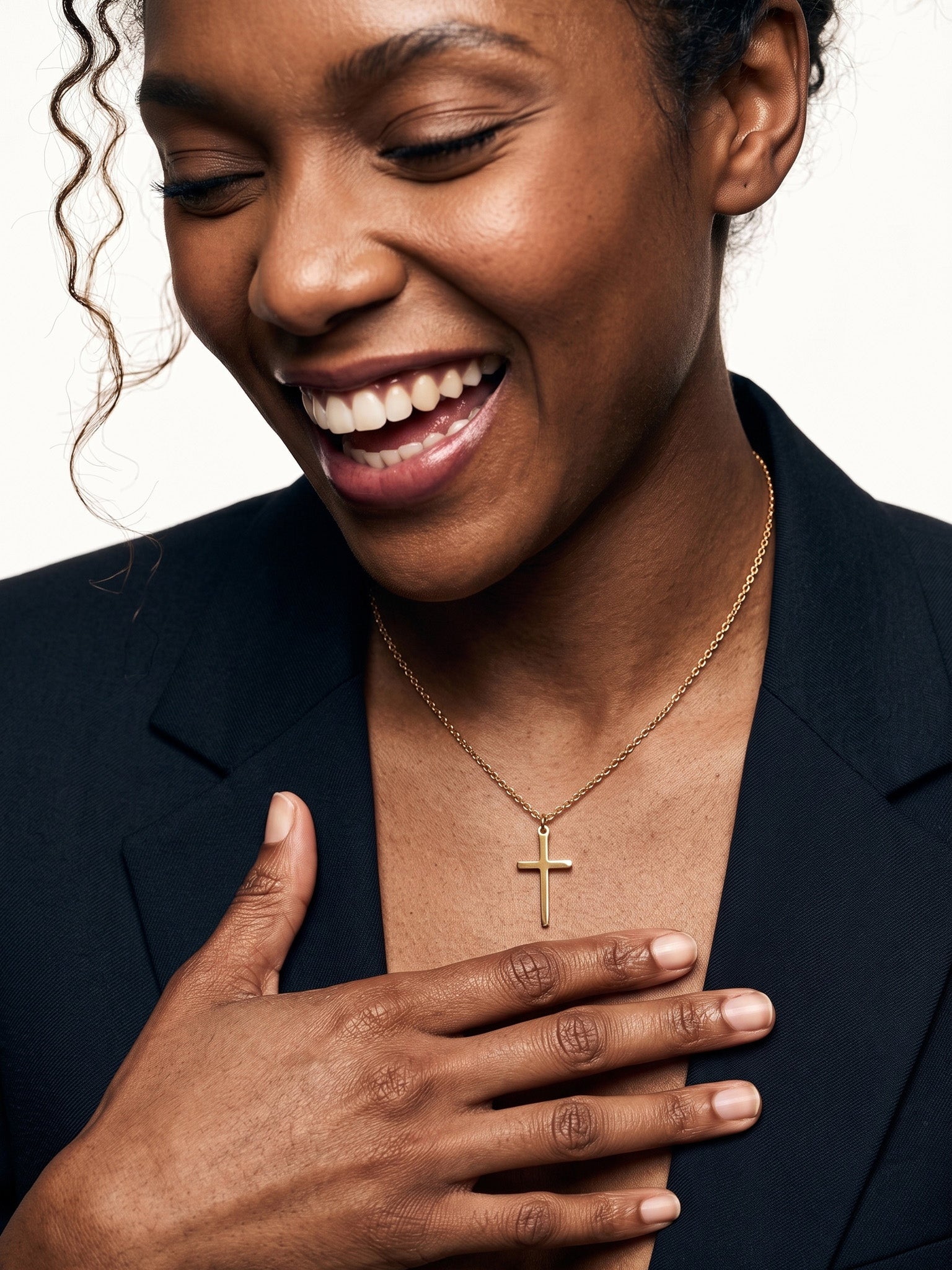 Steadfast Cross Necklace – worn on the neck, laughing, hand resting on the chest, gold, dark blazer, ivory studio background