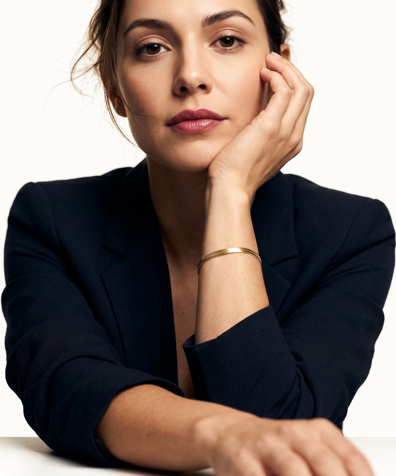 Still Waters Herringbone Bracelet – worn on the wrist, chin resting on the hand, seated at a table, gold, dark blazer, ivory studio background