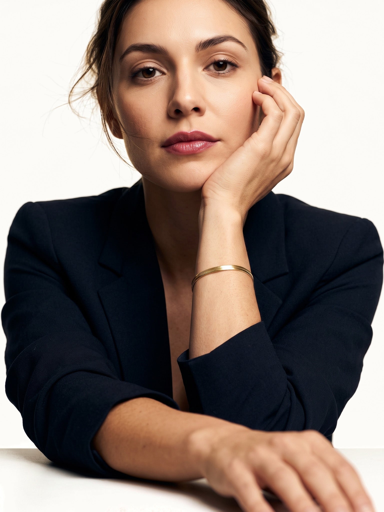Still Waters Herringbone Bracelet – worn on the wrist, chin resting on the hand, seated at a table, gold, dark blazer, ivory studio background