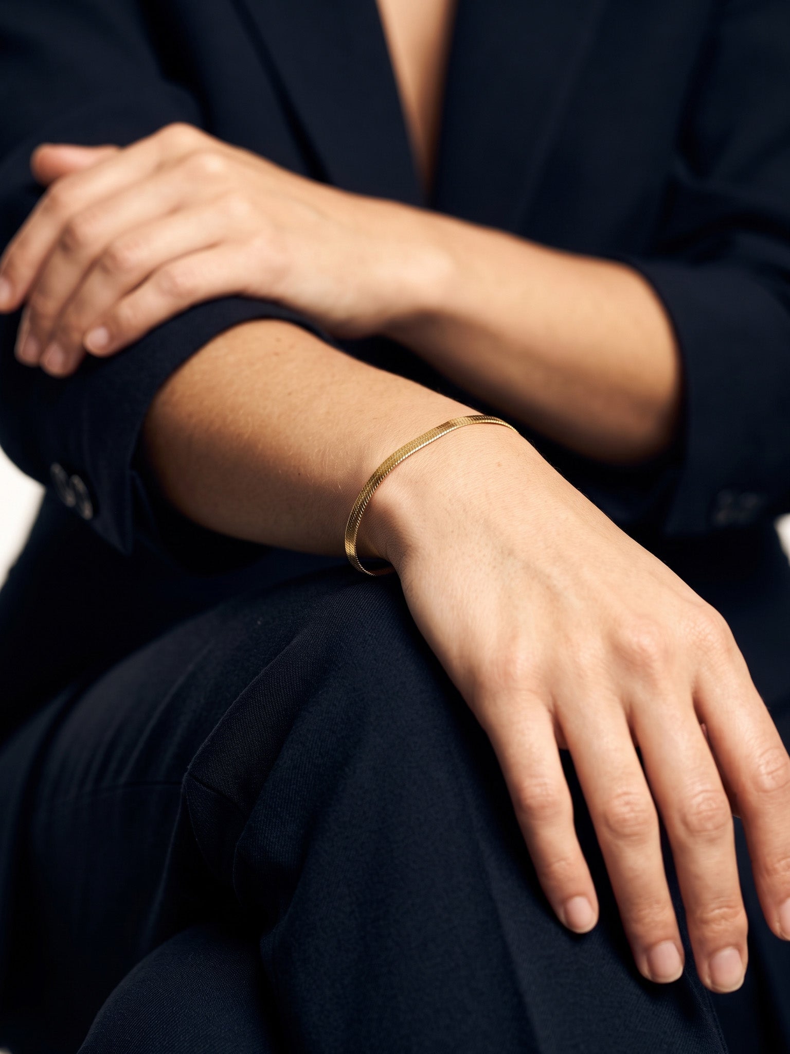 Still Waters Herringbone Bracelet – worn on the wrist, close-up, hand resting on the knee, gold, dark blazer, ivory studio background