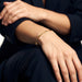 Still Waters Herringbone Bracelet – worn on the wrist, close-up, hand resting on the knee, gold, dark blazer, ivory studio background
