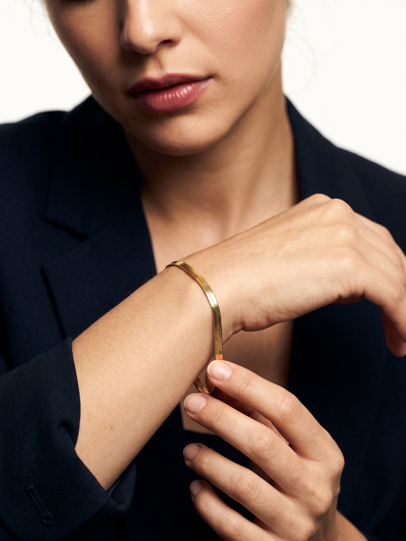 Still Waters Herringbone Bracelet – worn on the wrist, one hand adjusting the bracelet, gold, dark blazer, ivory studio background