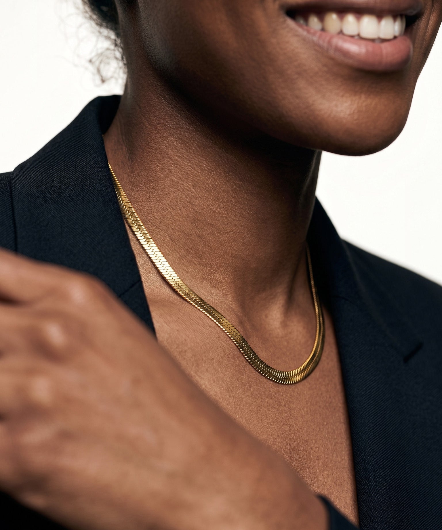 Sunday Best Herringbone Necklace – herringbone chain lying flat at the neck, smiling, hand at the shoulder, gold, black blazer, ivory studio background