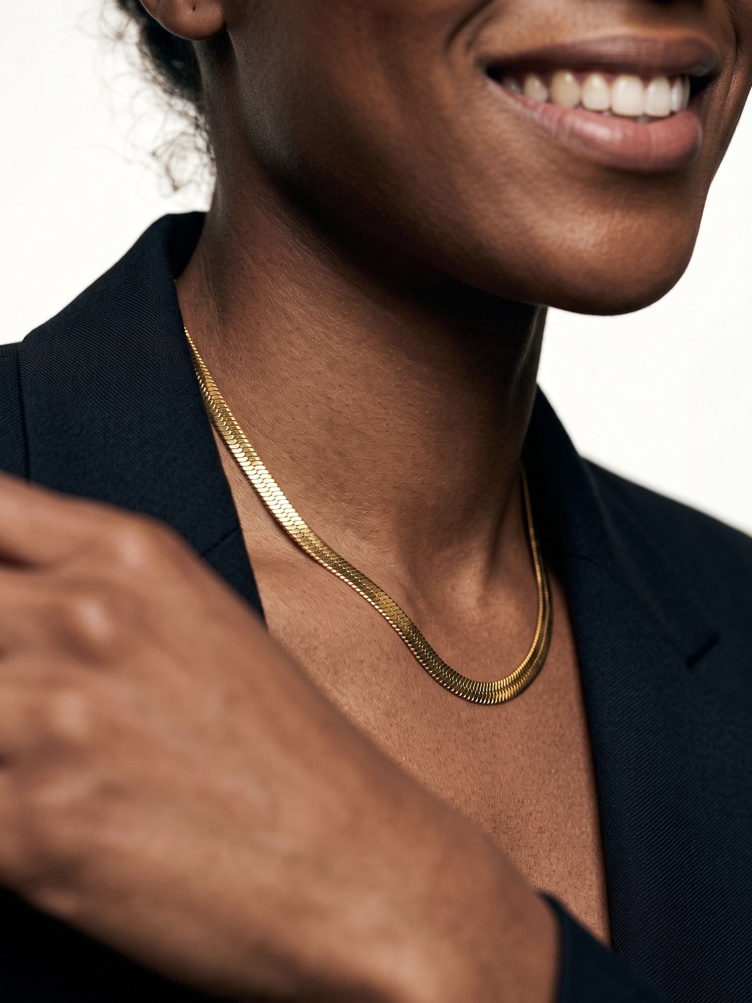 Fallen Herringbone Necklace – herringbone chain lying flat at the neck, smiling, hand at the shoulder, gold, black blazer, ivory studio background