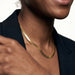 Fallen Herringbone Necklace – herringbone chain lying flat at the neck, smiling, hand at the shoulder, gold, black blazer, ivory studio background