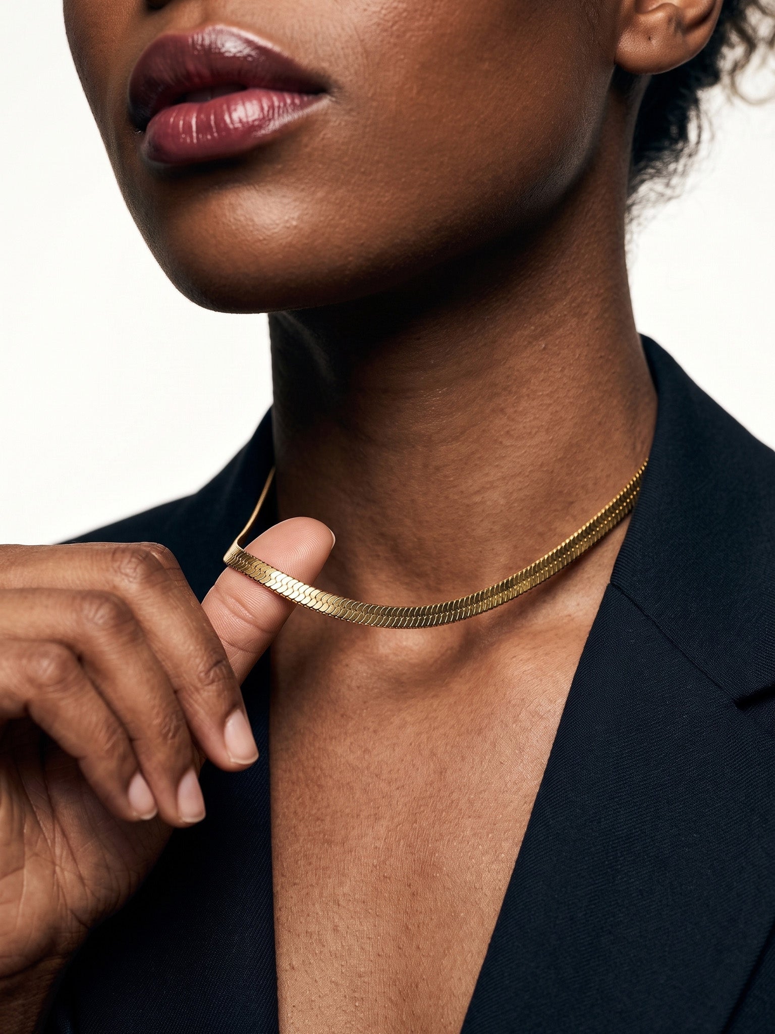 Fallen Herringbone Necklace – fingers lifting the herringbone chain at the neck, gold, black blazer, ivory studio background