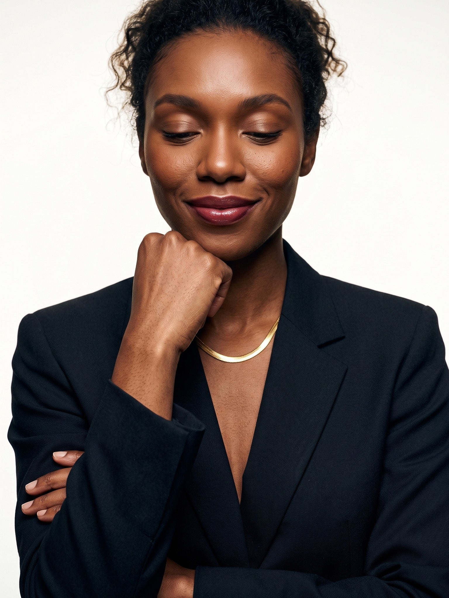Fallen Herringbone Necklace – worn at the collarbone, eyes closed with a fist at the chin, gold, black blazer, ivory studio background