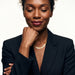 Fallen Herringbone Necklace – worn at the collarbone, eyes closed with a fist at the chin, gold, black blazer, ivory studio background