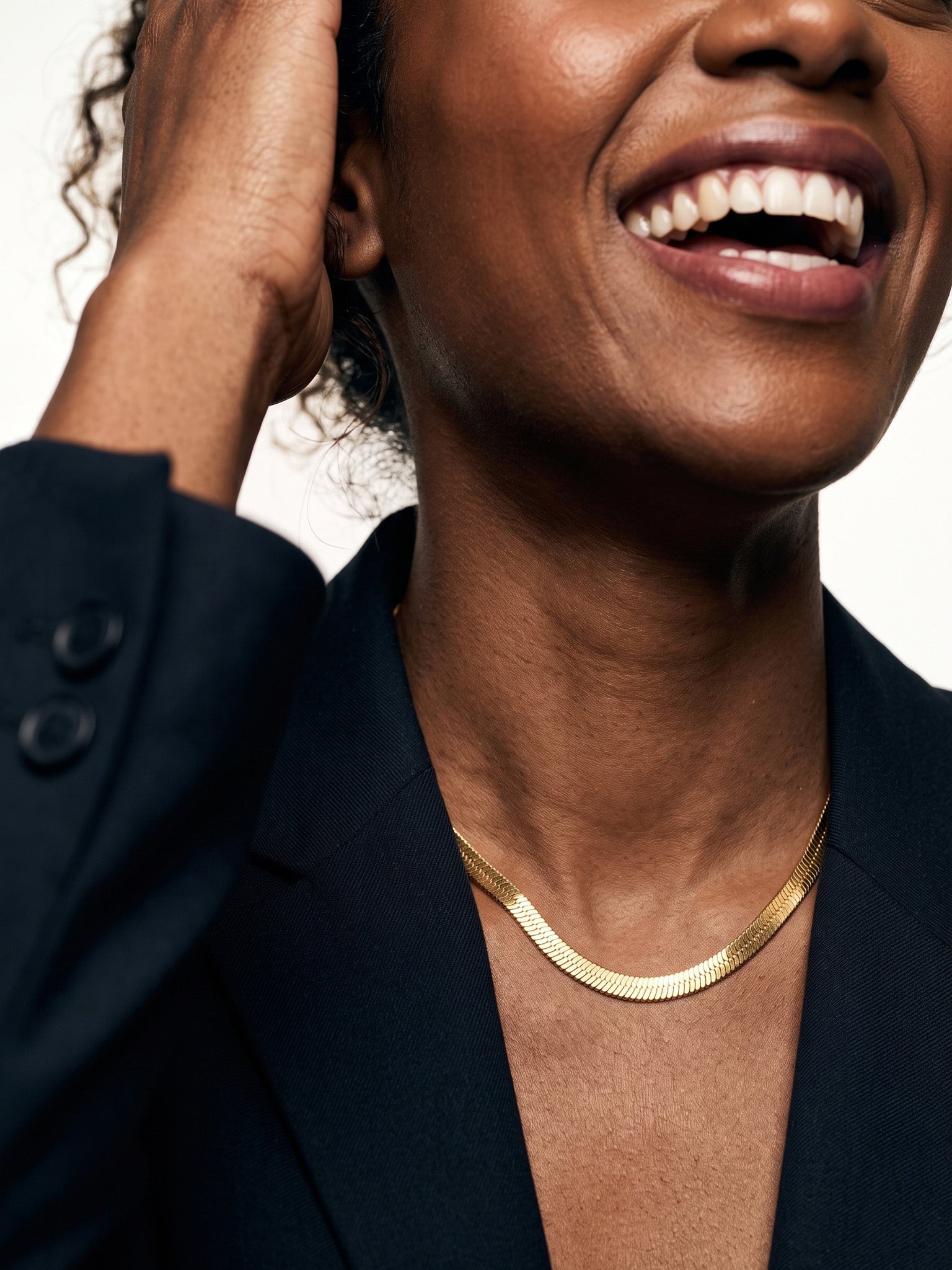 Fallen Herringbone Necklace – close crop of the herringbone chain at the neck, laughing, hand in hair, gold, black blazer, ivory studio background