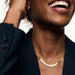 Fallen Herringbone Necklace – close crop of the herringbone chain at the neck, laughing, hand in hair, gold, black blazer, ivory studio background