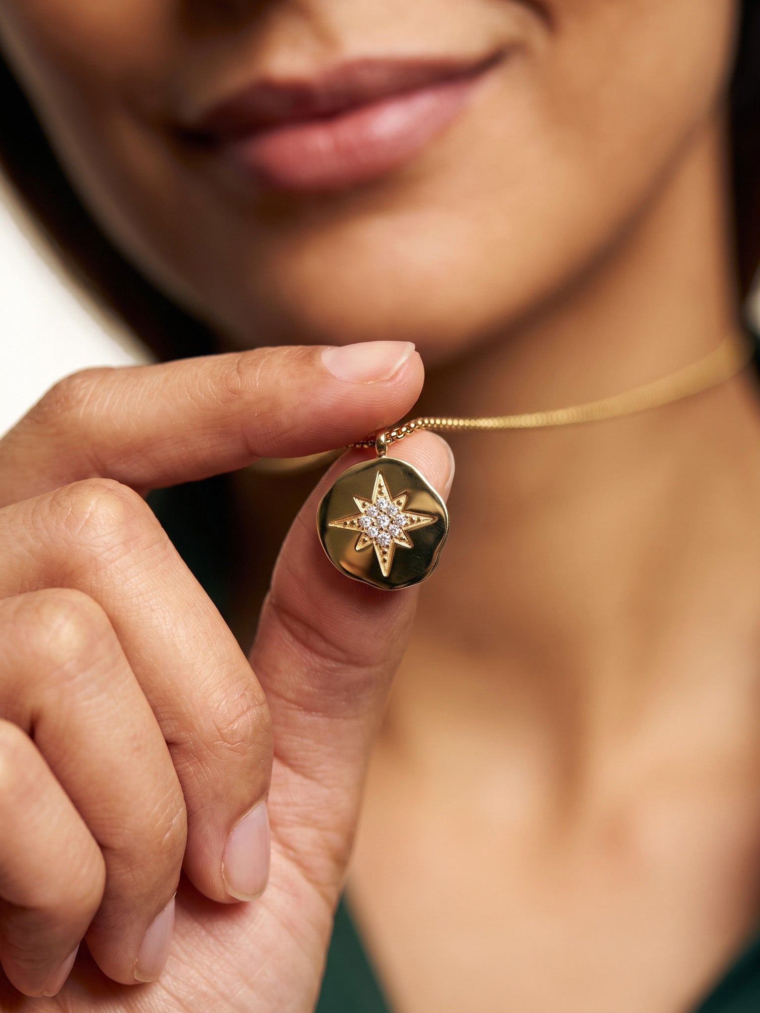 True North Necklace – star-compass medallion held between finger and thumb, close crop, gold, ivory studio background