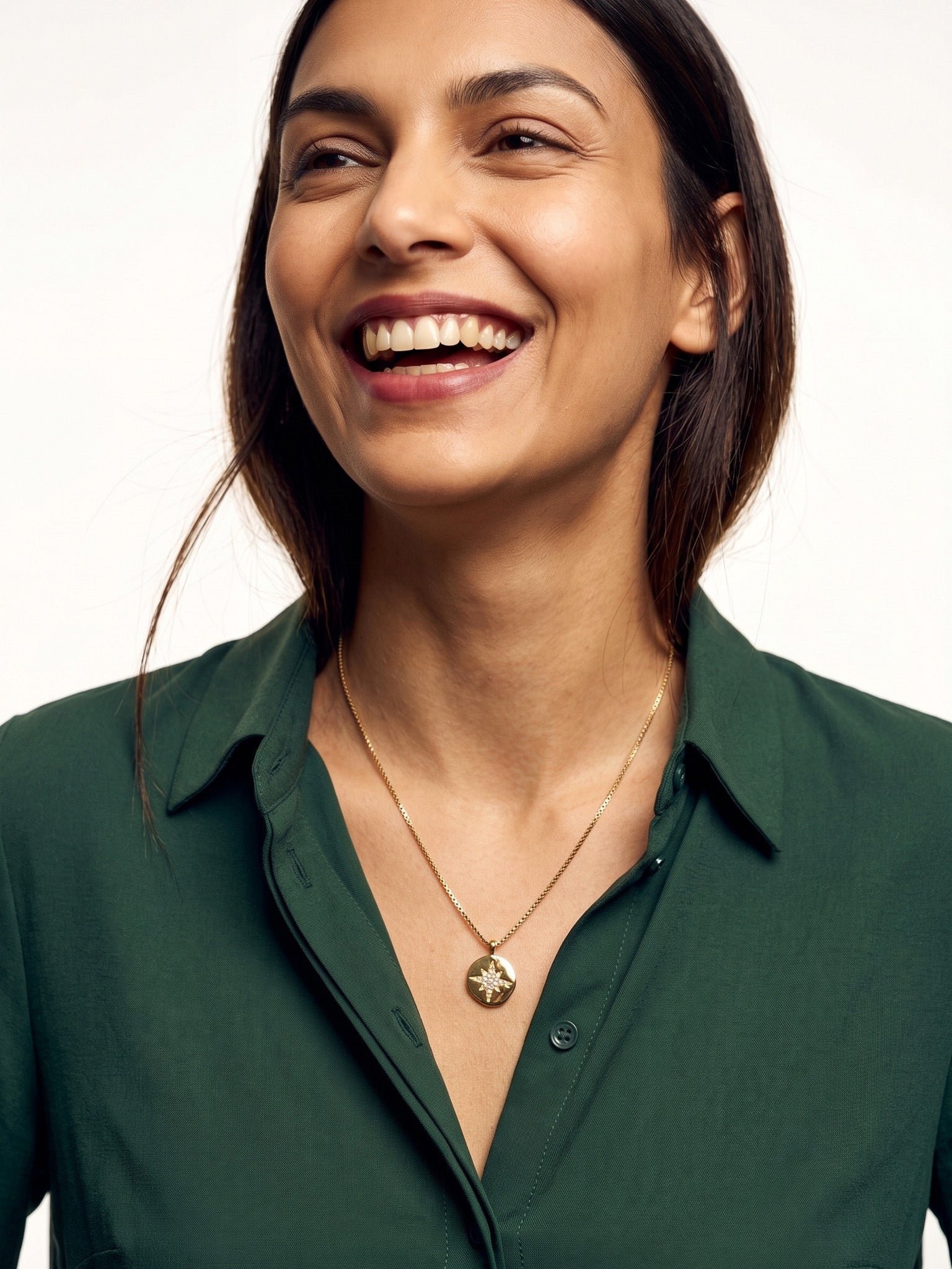 True North Necklace – worn at the chest, laughing, gold, deep green shirt, ivory studio background